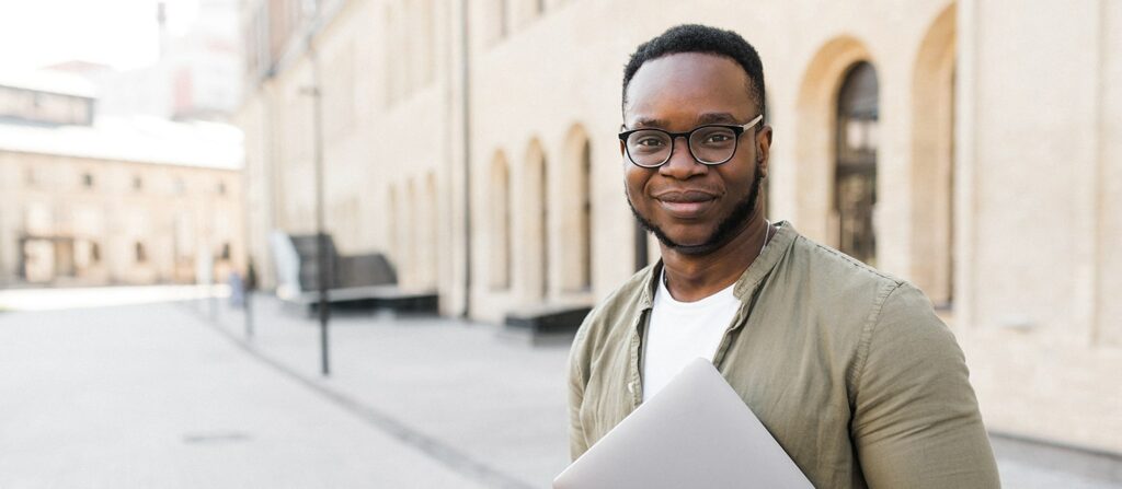 Jeune homme de face tenant un ordinateur sur sa poitrine devant une université dans al rue